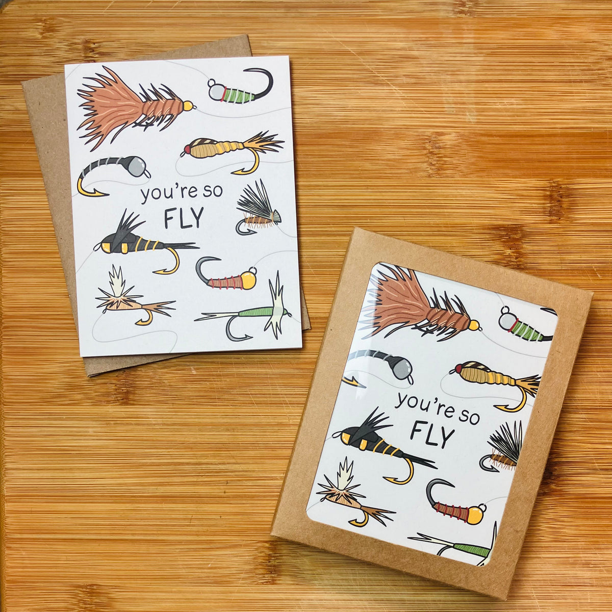 A card and stationery set featuring illustrations of fly fishing hooks and the phrase “you’re so FLY,” photographed against a wood surface.