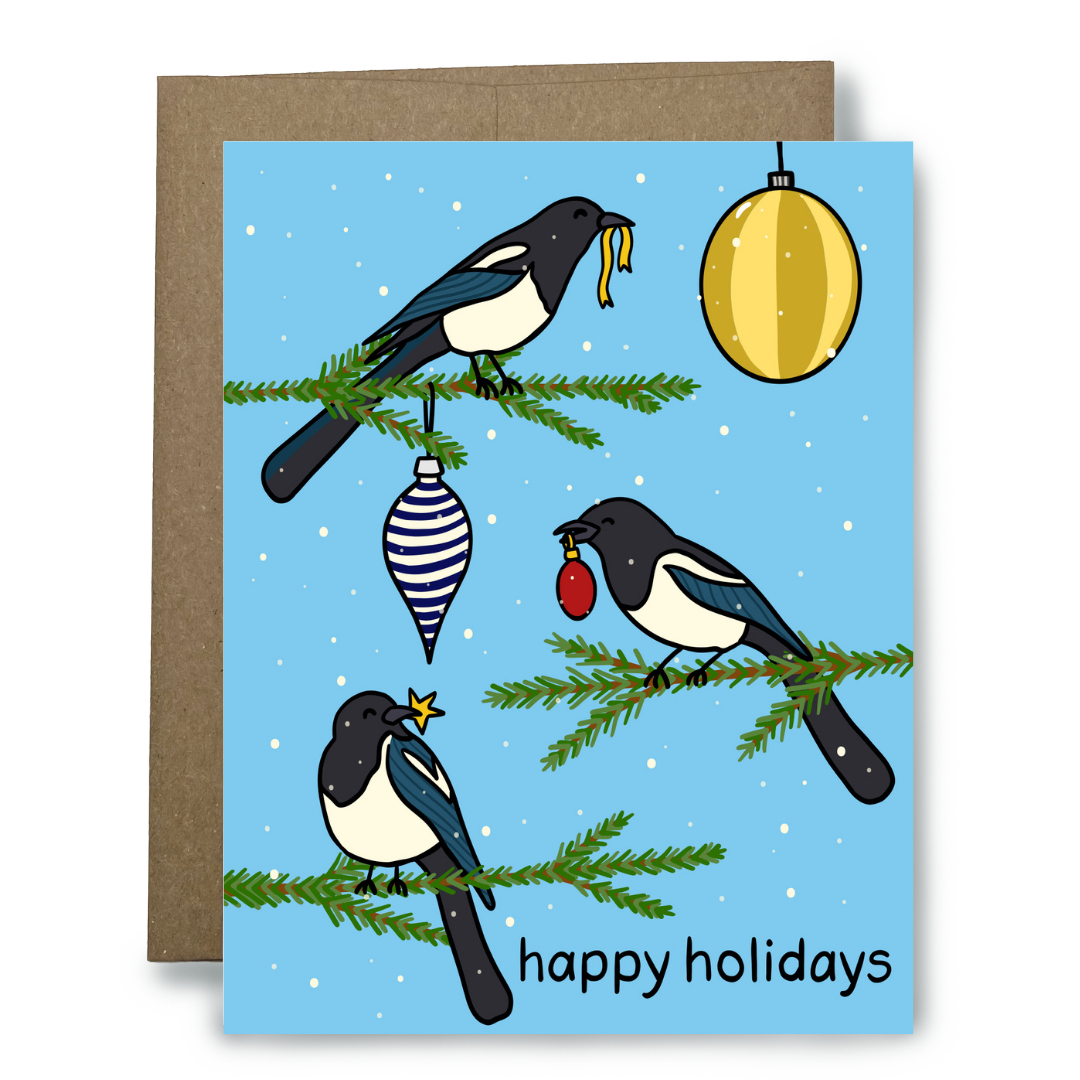 A holiday greeting card with a brown envelope behind it. The card features an illustration of three magpies perched on Christmas tree branches against a light blue snowy background, with hanging ornaments and the text 'happy holidays' at the bottom.