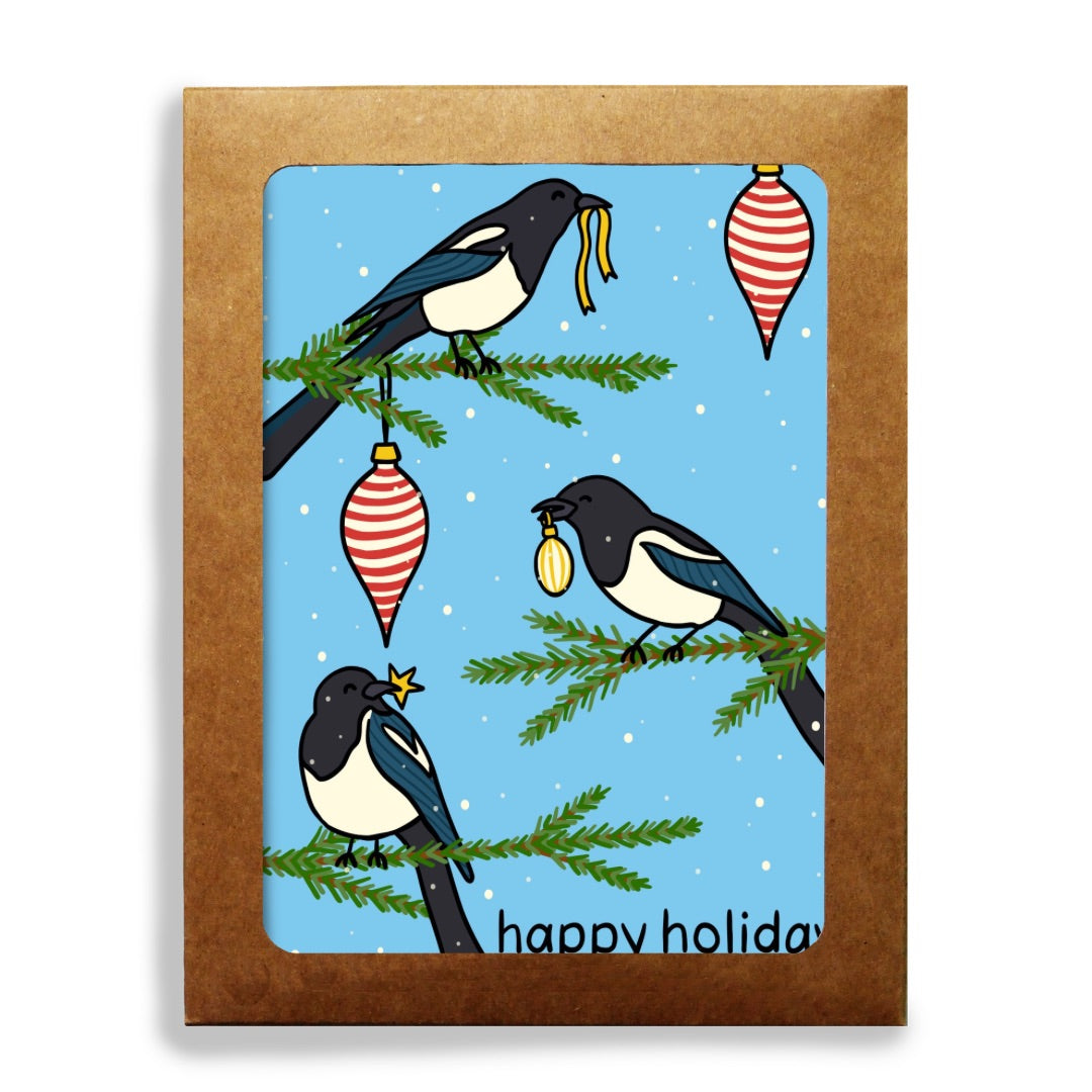 A rectangular stationery set and a blue illustrated card showing three magpie birds on evergreen branches with hanging red-and-white ornaments and the text 'happy holiday' at the bottom.
