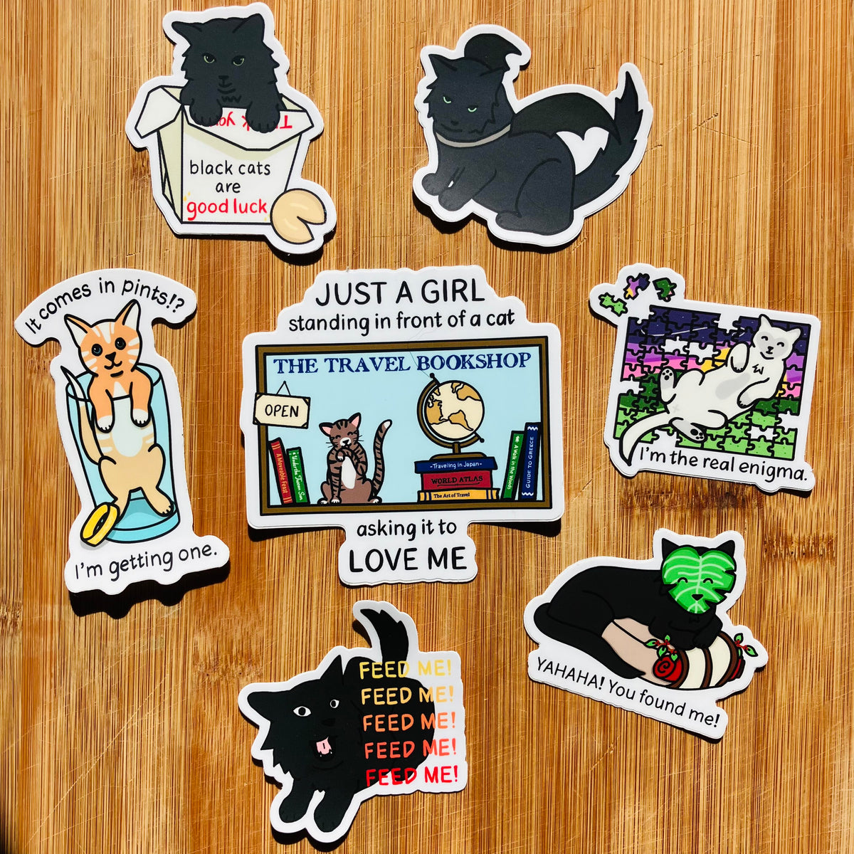 A set of seven die-cut cartoon cat stickers displayed on a light brown wooden background. The stickers feature various humorous and playful cat-themed designs
