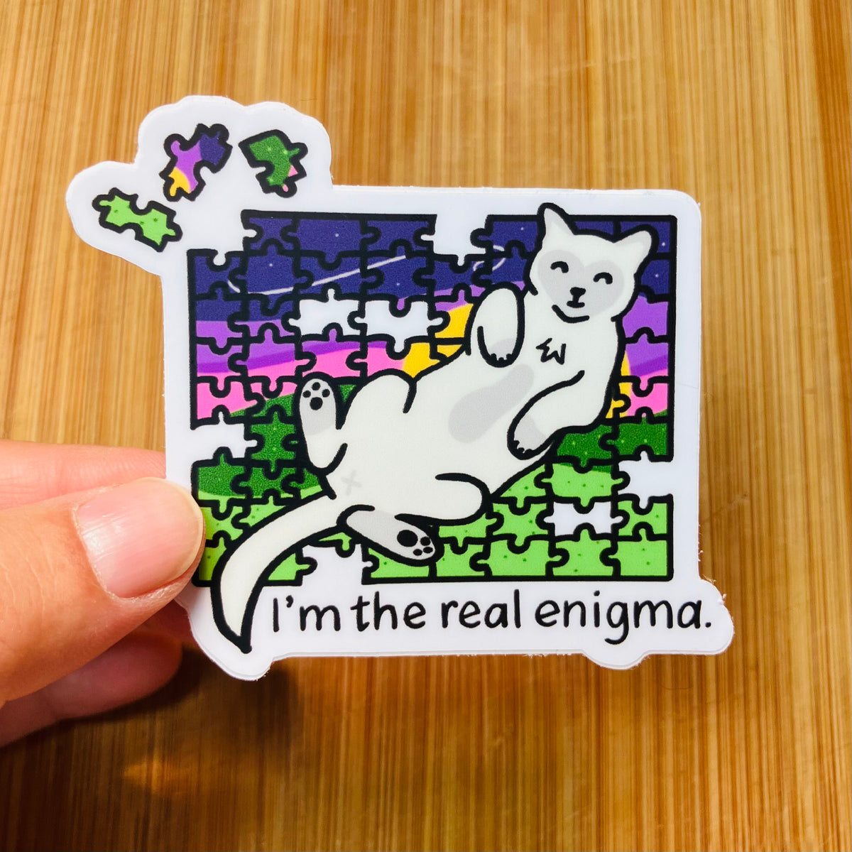 A hand holds a die-cut sticker featuring a white cat lying on a colorful jigsaw puzzle with the text 'I'm the real enigma.' The sticker is photographed against a light wood surface.