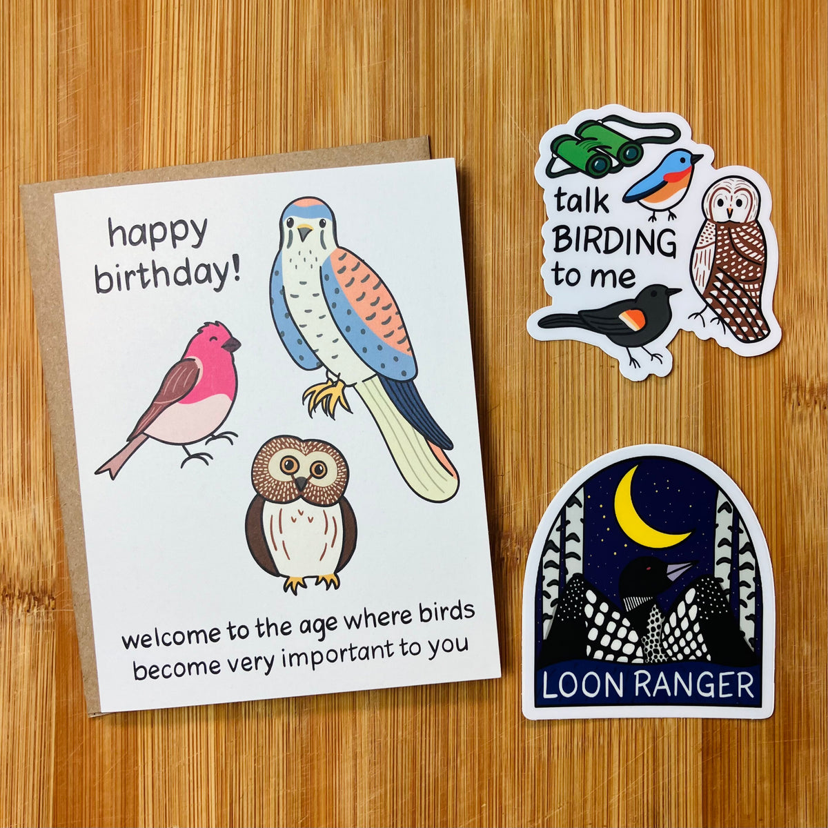A birthday card with bird illustrations and the text 'happy birthday! welcome to the age where birds become very important to you' is shown on a wood surface alongside two bird-themed stickers.