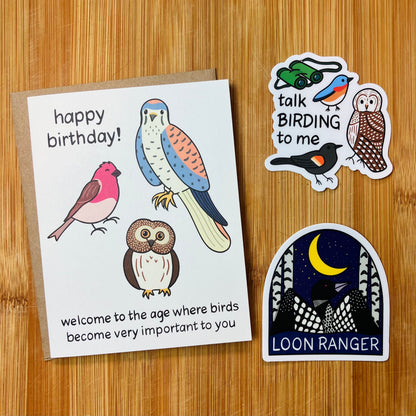 A birthday card with bird illustrations and the text 'happy birthday! welcome to the age where birds become very important to you' is shown on a wood surface alongside two bird-themed stickers.