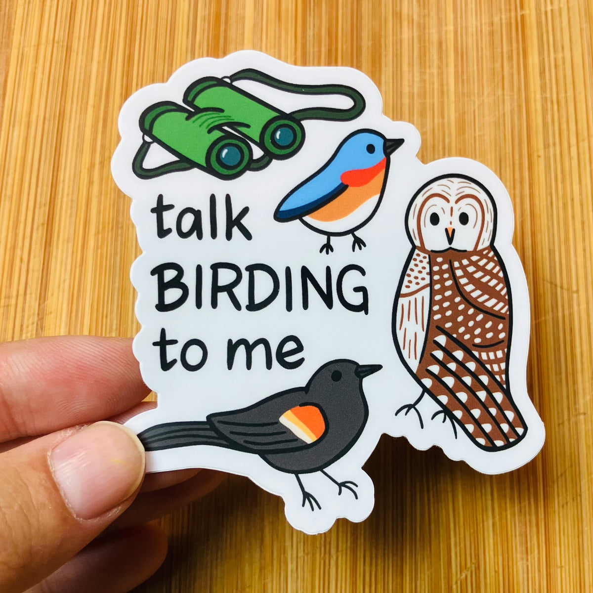A hand holds a die-cut sticker featuring the phrase 'talk BIRDING to me' with illustrated birds and binoculars, placed on a light wood surface.