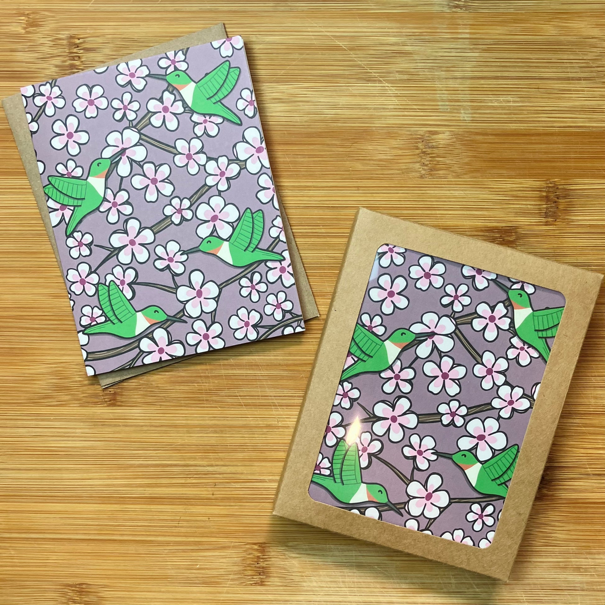 A greeting card and a stationery set with a printed cherry blossom and hummingbird design, displayed on a light wood surface.