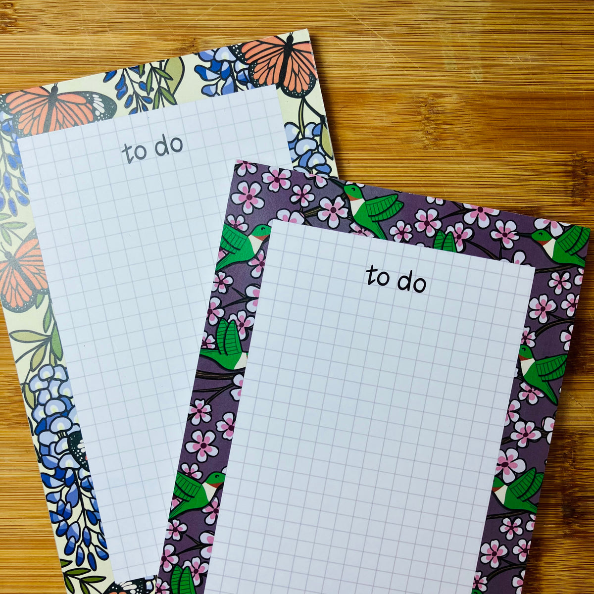 Two floral 'to do' grid notepads on a wooden surface, one with a wisteria and butterfly border and the other with a cherry blossom and hummingbird border.