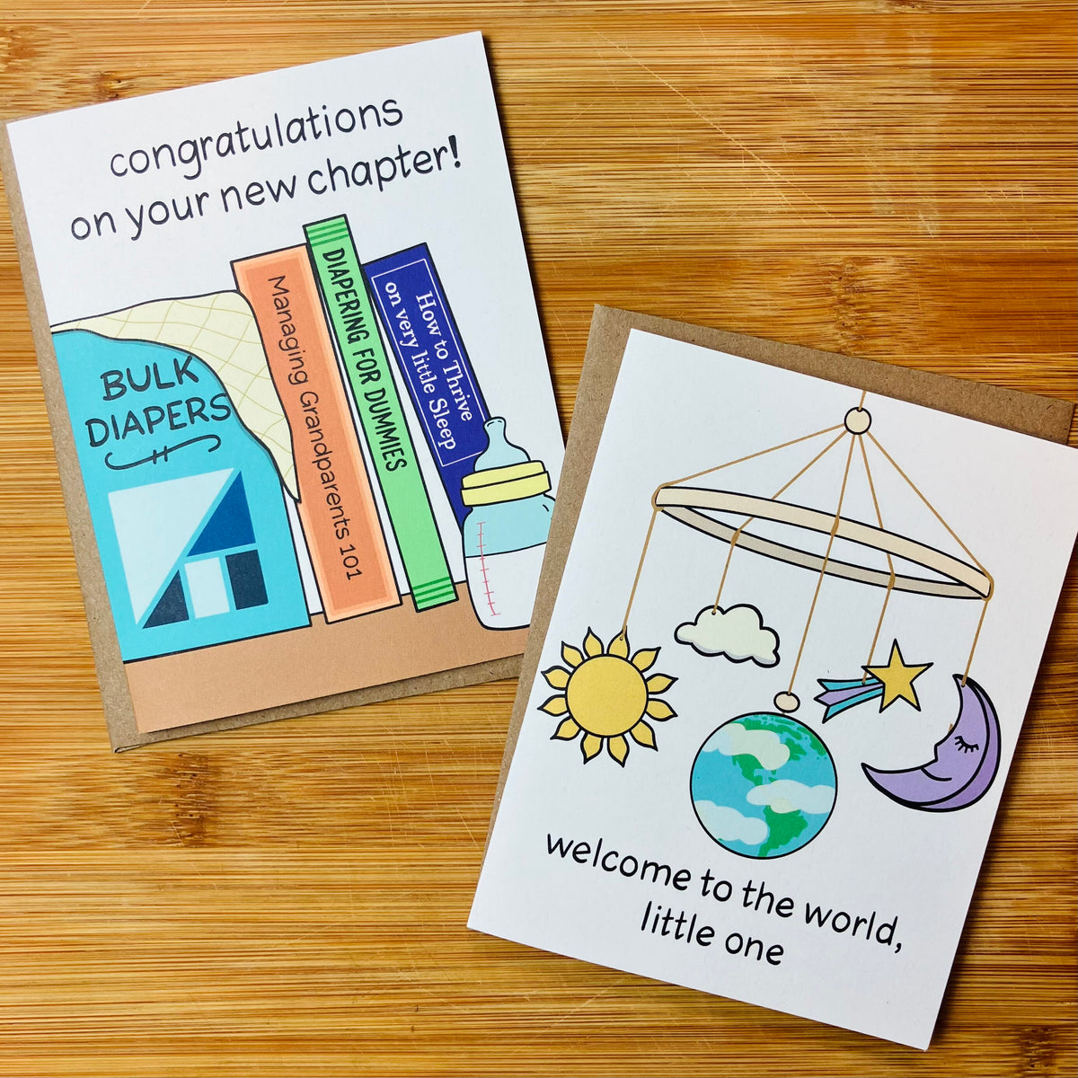 Two baby themed greeting cards displayed on a wooden tabletop. One card reads 'Congratulations on your new chapter!' with colorful book illustrations, and the other says 'Welcome to the world, little one' with a moon, sun, cloud, and Earth baby mobile illustration.
