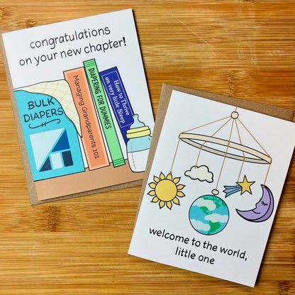Two baby themed greeting cards displayed on a wooden tabletop. One card reads 'Congratulations on your new chapter!' with colorful book illustrations, and the other says 'Welcome to the world, little one' with a moon, sun, cloud, and Earth baby mobile illustration.