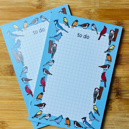 Two grid to-do lists with bird illustrations around the border, placed on a wooden surface.