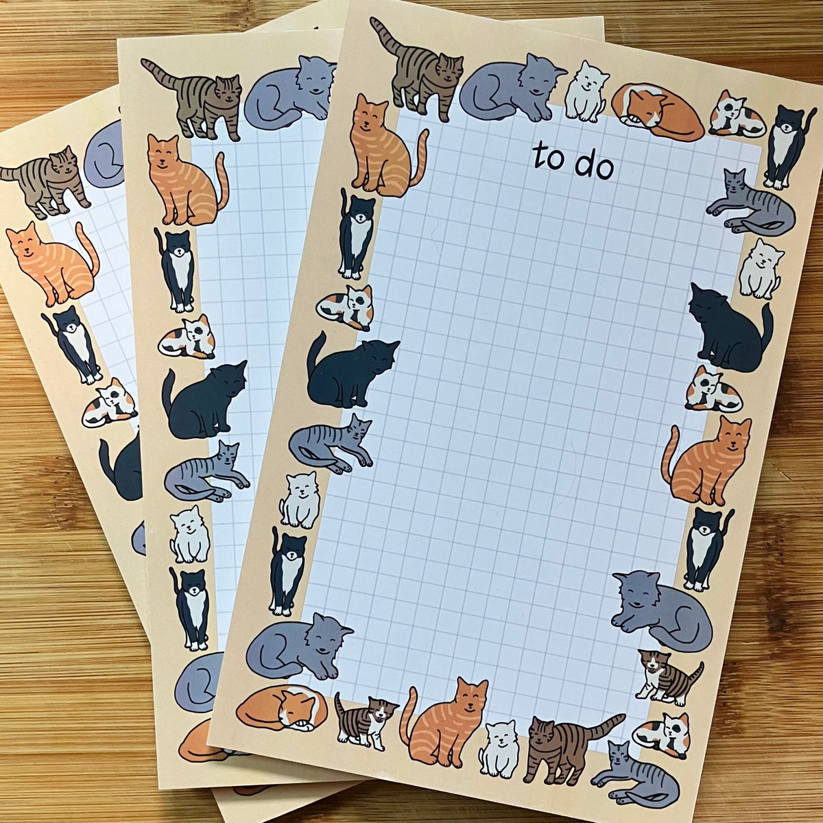 A set of cat-themed to-do list pads with a border of illustrated cats in various colors, displayed on a wooden surface.