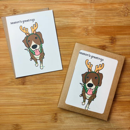 A greeting card and stationery setare arranged on a wooden surface. Each card has a white front with a cartoon dog wearing antlers, a green wreath, and the text 'season's greetings'.
