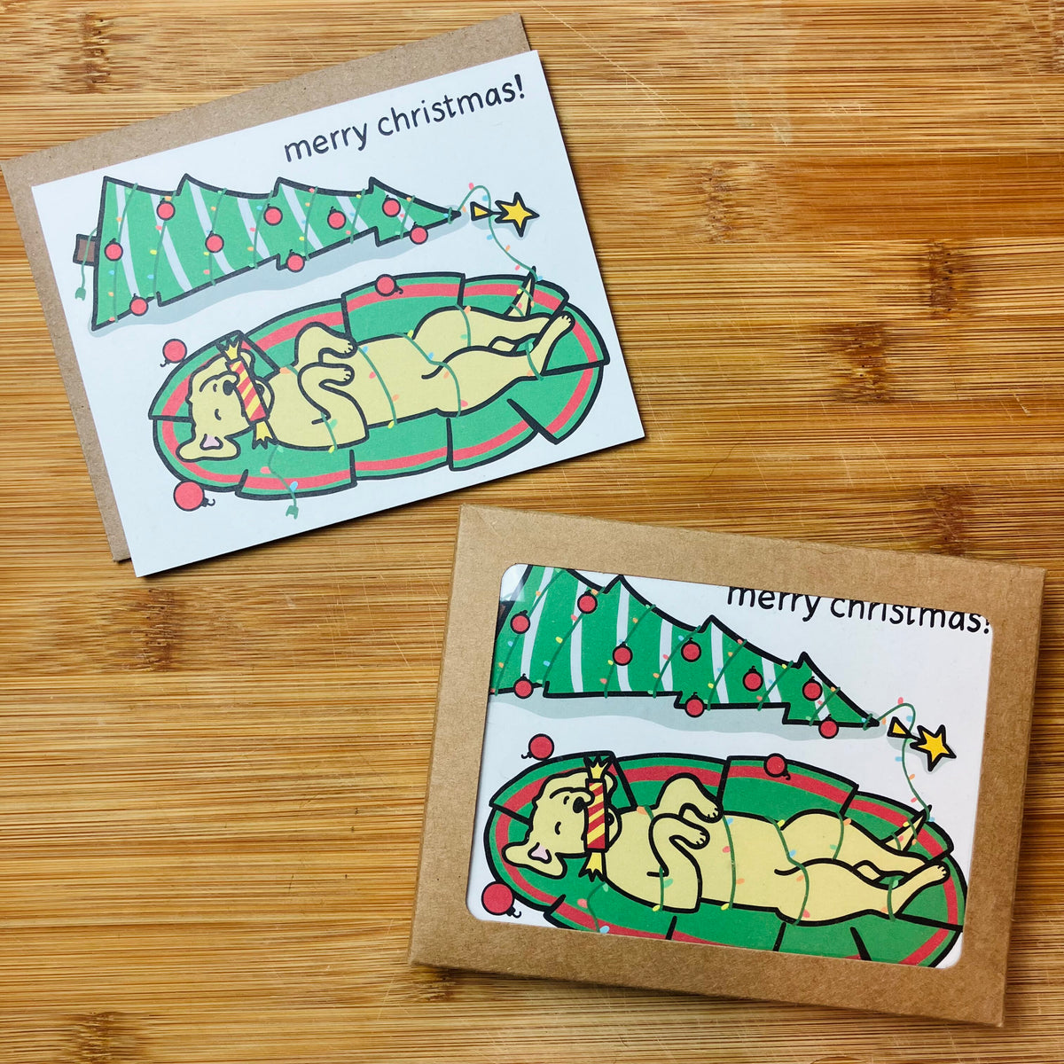 Christmas card and stationery set, both featuring a dog wrapped in Christmas lights and a fallen down Christmas tree in the background, with 'merry christmas!' text, photographed against a wood backdrop.