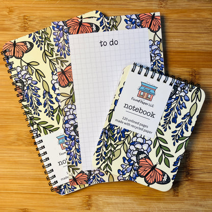 Two notebooks and a to do pad with butterfly and wisteria design, photographed against warm wood background