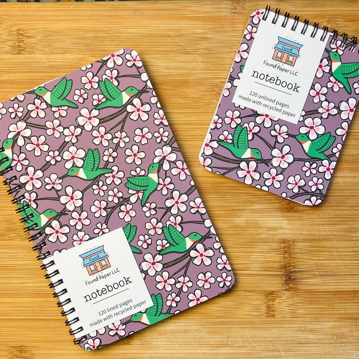 Two notebooks with designs of green hummingbirds and cherry blossoms, photographed against a warm wood background