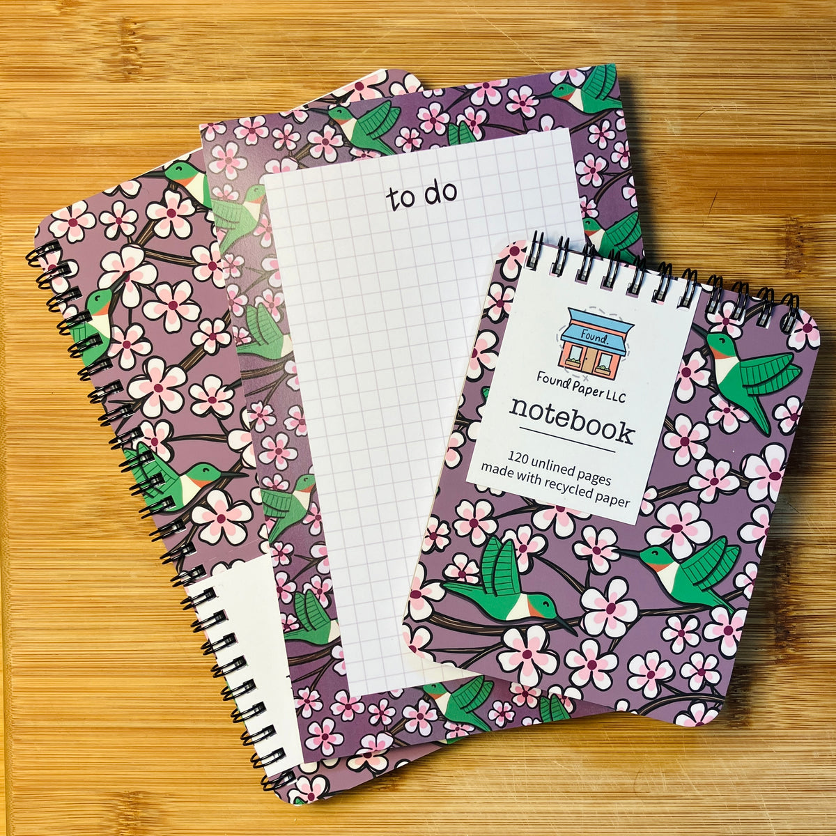 Two notebooks and a to-do pad with designs of green hummingbirds and cherry blossoms, photographed against a warm wood background