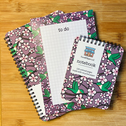 Two notebooks and a to-do pad with designs of green hummingbirds and cherry blossoms, photographed against a warm wood background