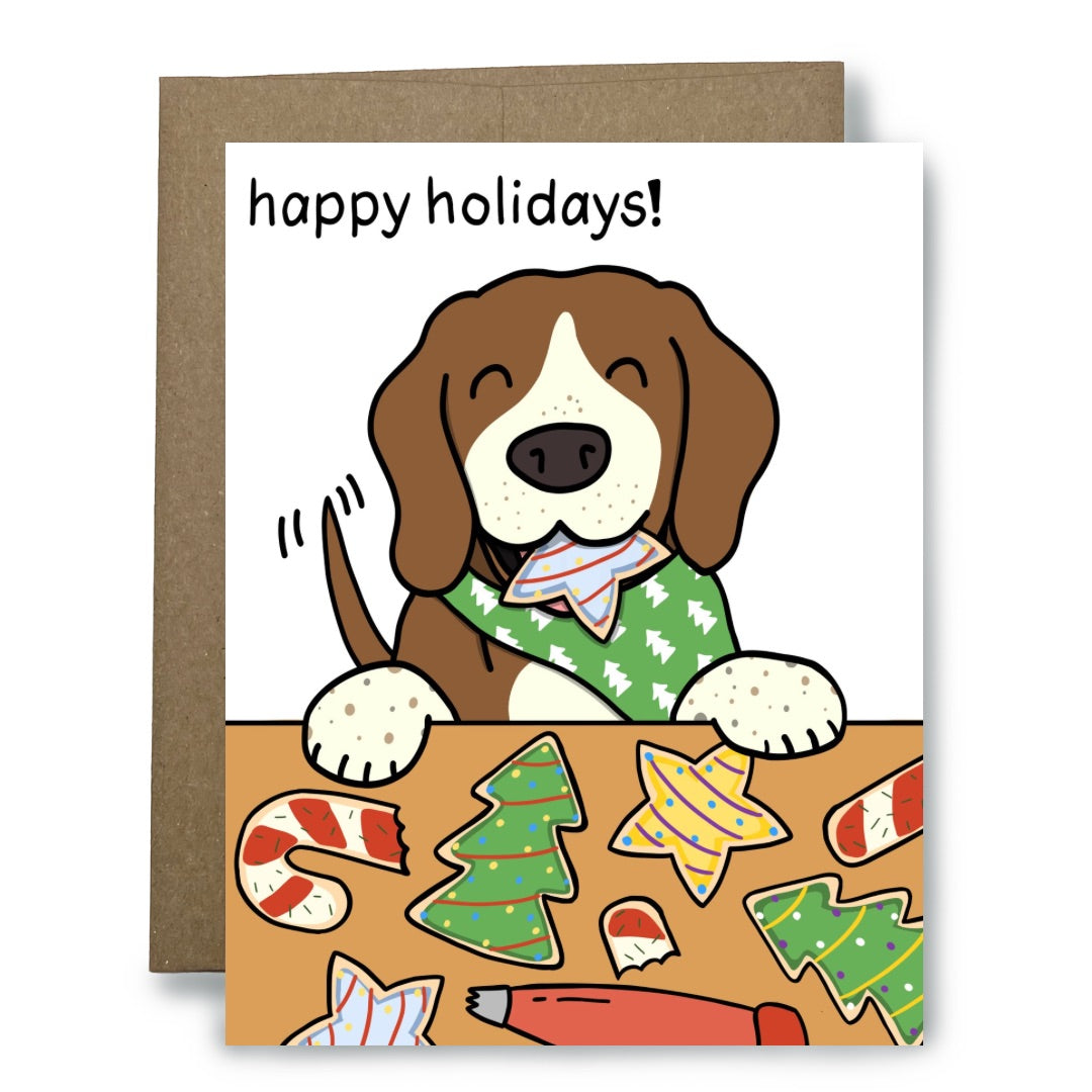 A holiday greeting card with a brown envelope behind it. The card features a cartoon beagle wearing a green Christmas bandanna and holding a cookie, with the text 'happy holidays!' above.